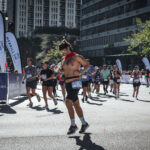 A group of runners participates in the Chicago Marathon on a sunny day in downtown Chicago, with tall buildings, race banners, and spectators lining the street. One runner in the foreground is shirtless, wearing sunglasses, a headband, a red scarf, and themed running socks while mid-stride.