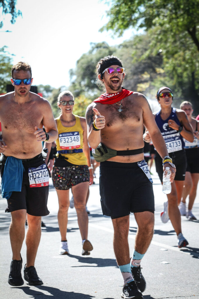 A group of runners participating in the Chicago Marathon moves along the course under bright daylight. One runner in the foreground smiles, points playfully toward the camera, and carries a water bottle while wearing sunglasses, a red neck gaiter, and race bib G42180. Other runners wearing race bibs and athletic gear surround him on the road.