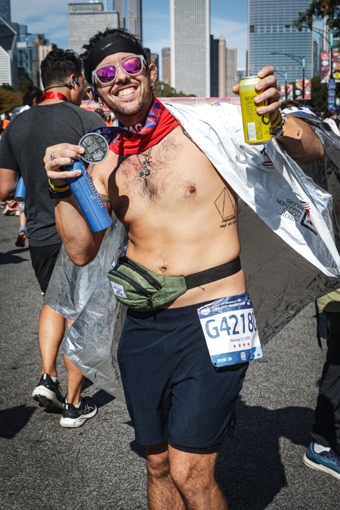 A marathon finisher smiles widely while holding his medal in one hand and two recovery beverages in the other. He wears reflective sunglasses, a red neck gaiter, a heat-retention blanket, a green running belt, and race bib G42180. Tall Chicago skyscrapers rise behind him as runners continue to cross the finish area.